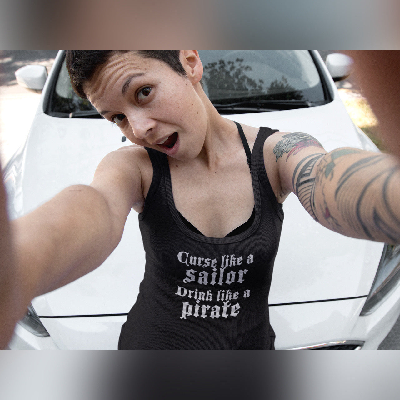 Woman with short dark hair takes a selfie. She wears a black tank top with the words curse like a sailor drink like a pirate in white.