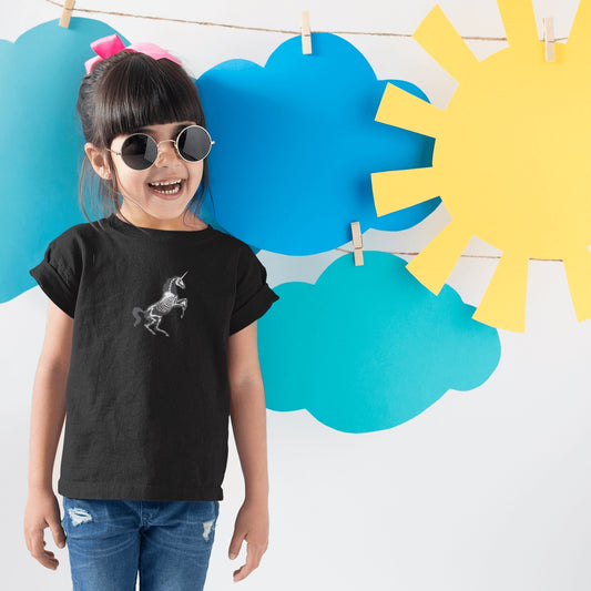 Brunette girl with sunglasses stands in a black unicorn skeleton youth t-shirt in front of cutout clouds and sun